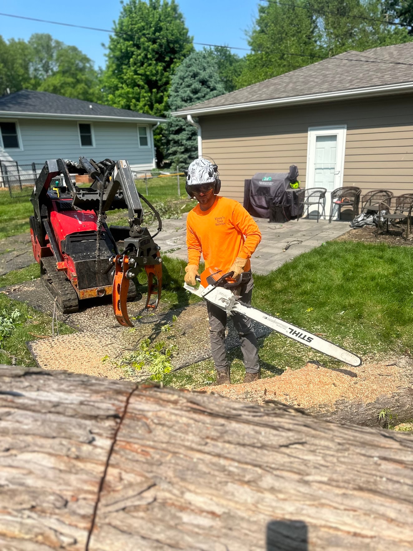 Vinny's Tree Service crew member in Vinny's orange shirt operating chainsaw with skid steer on job site